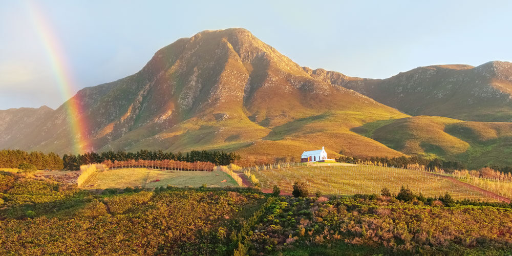 Spektakulärer Blick auf ein Bergmassiv in Südafrika vor dem ein Regenbogen direkt auf ein mit Wein angebautes Feld scheint