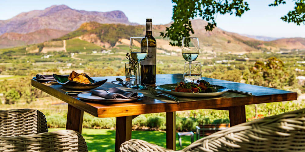 Ein mit Weingläsern und Weinflasche gedeckter Holztisch steht im Baumschatten vor einer schönen Bergkulisse in Südafrika
