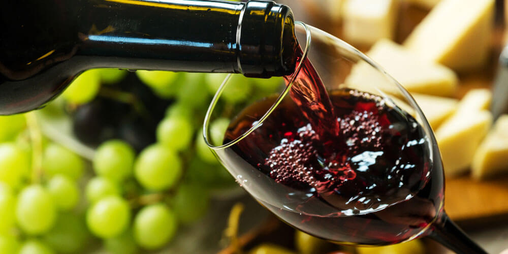 Tiefroter Wein aus der Flasche wird in ein schräg gehaltenes Weinglas geschenkt, dahinter liegen Weintrauben und Käsewürfel