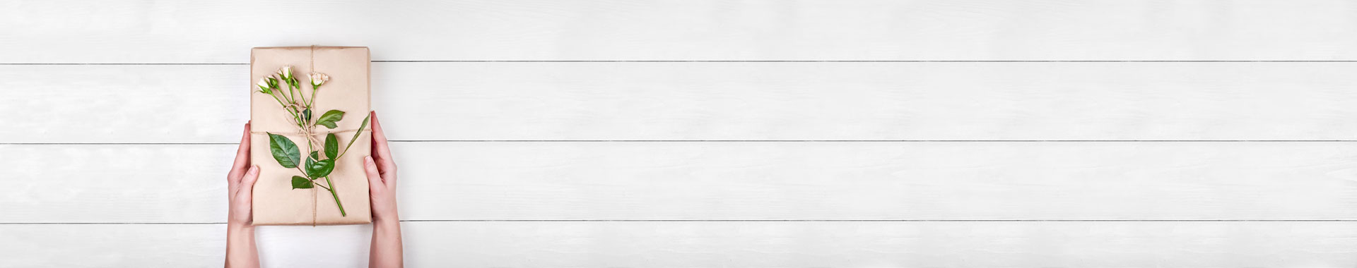 Ein schön verpacktes Geschenk verziert mit einem Rosenstiel wird von zwei Händen vor einer Holzplatte gehalten